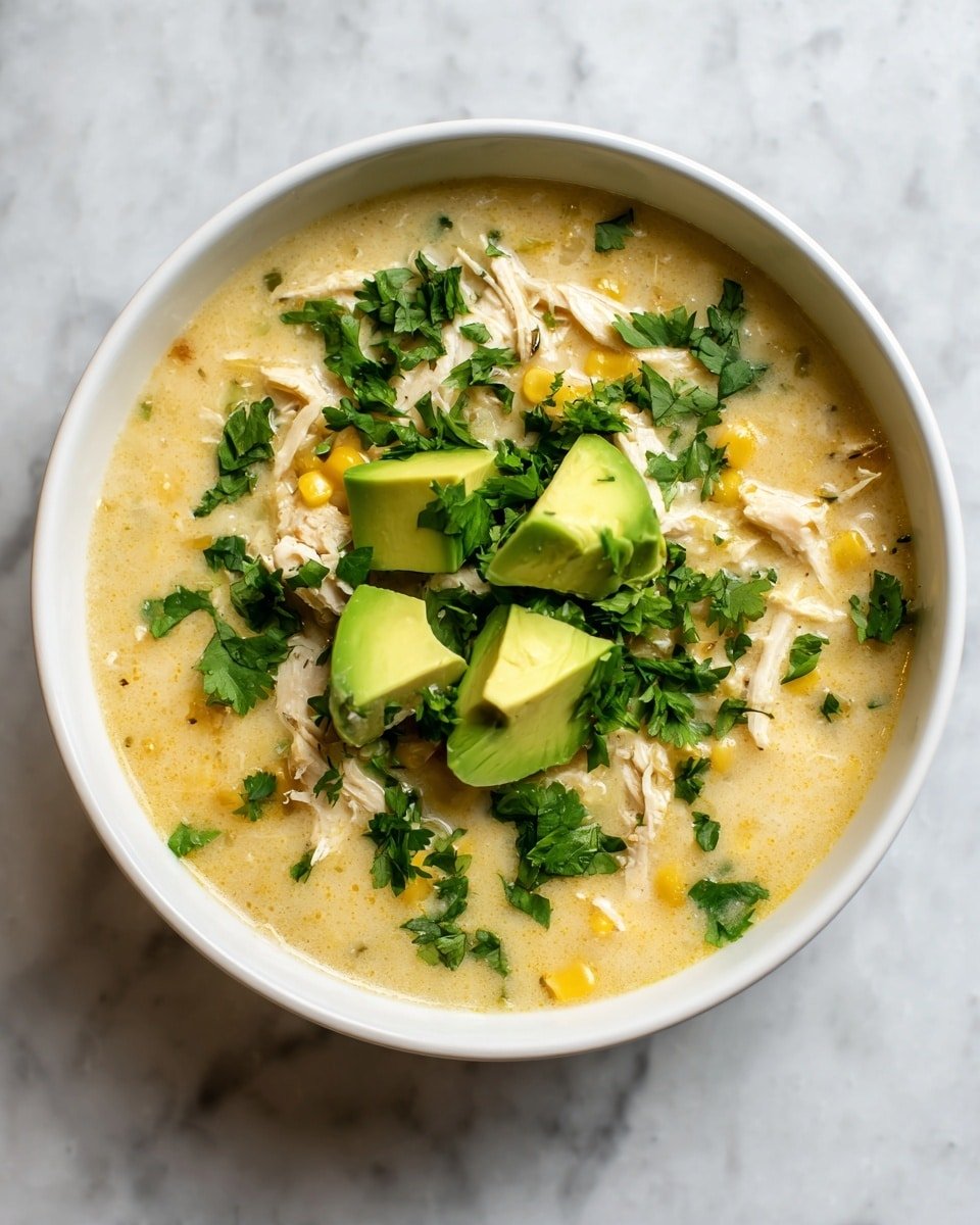 A top view of a white bowl filled with creamy soup that has a light yellow color and a smooth texture mixed with small pieces of corn and shredded white chicken. On top, there are five chunks of bright green avocado placed in the center, surrounded by scattered fresh green cilantro leaves. The bowl is placed on a white marbled surface, making the colors of the soup and toppings stand out clearly. photo taken with an iphone --ar 4:5 --v 7 — Keto White Chicken Chili, low-carb chicken chili, healthy white chicken chili, creamy keto chili, gluten-free chicken chili