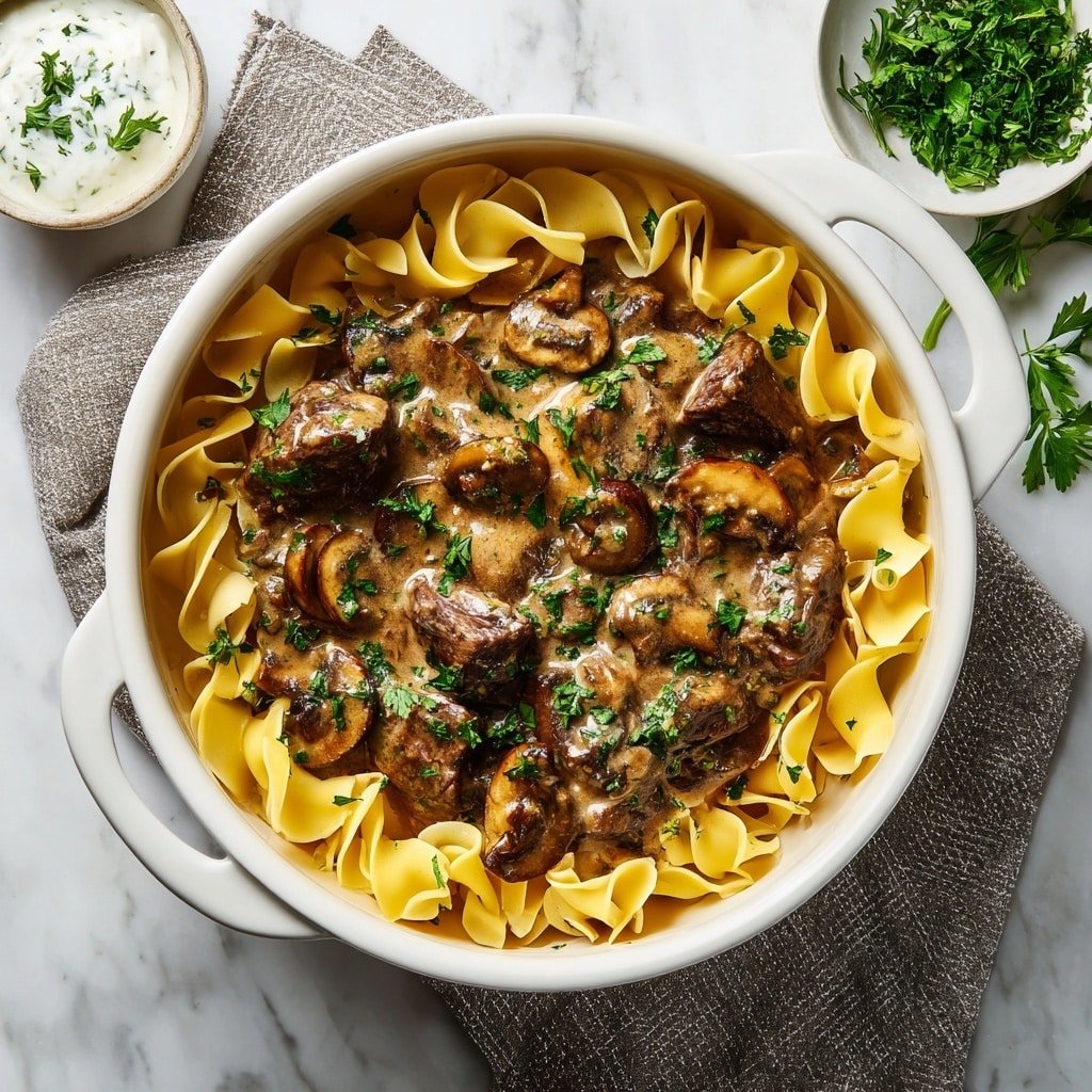 A round white pot filled with a rich brown stew made of beef chunks and sliced mushrooms in a creamy sauce, surrounded by wide yellow egg noodles placed around the edges inside the pot. The beef pieces are medium-sized and scattered in the center with some fresh green parsley sprinkled on top for color. The pot sits on a white marbled surface, with a textured gray cloth and a small white bowl of white sauce nearby, and green parsley sprigs lying to the upper right. Photo taken with an iphone --ar 1:1 --v 7 — One-Pot Beef Stroganoff, beef stroganoff dinner ideas, easy beef stroganoff recipe, creamy beef mushroom skillet, quick weeknight beef casserole