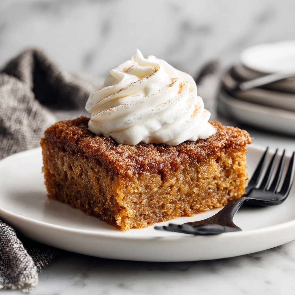 The image shows a single piece of baked dessert with a rough texture and brown color, topped with a swirl of white whipped cream in the center. The dessert looks dense with visible cinnamon and sugar coating, and it is placed on a plain white round plate. The plate rests on a white marbled surface with a gray cloth and black fork nearby, adding subtle texture to the background. photo taken with an iphone --ar 1:1 --v 7 — Pumpkin French Toast Bake, fall breakfast casserole, pumpkin breakfast ideas, easy autumn brunch recipes, cozy holiday breakfast